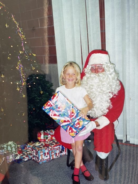 Courtney Shondel as a little girl, smiling with a wrapped holiday gift beside Santa